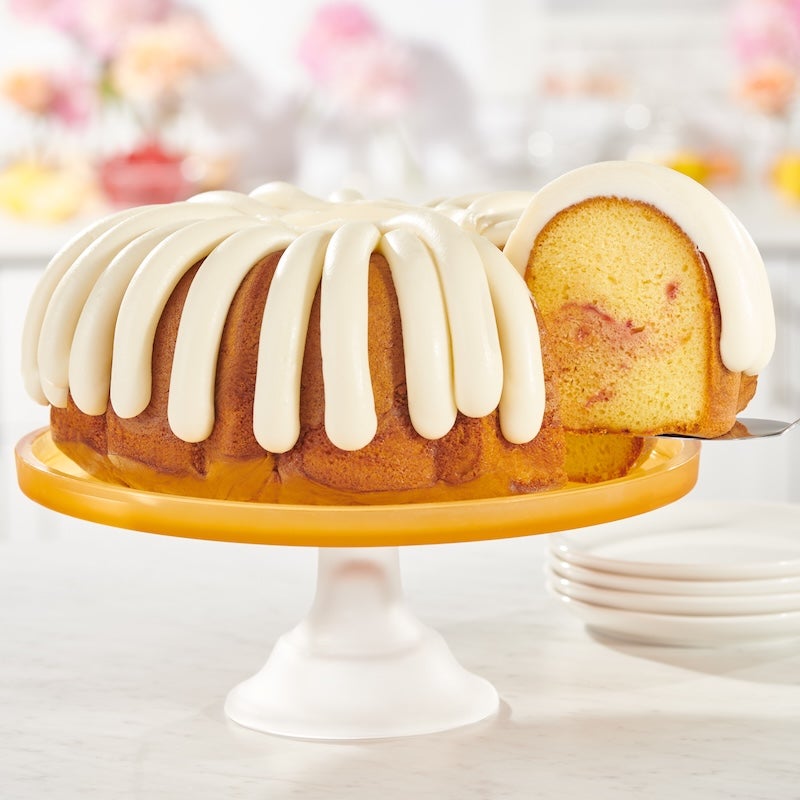 Lemon Raspberry Bundt cake crowned with signature cream cheese frosting with a slice showing the raspberry swirl in the middle of the cake - Shop Bundt Cakes