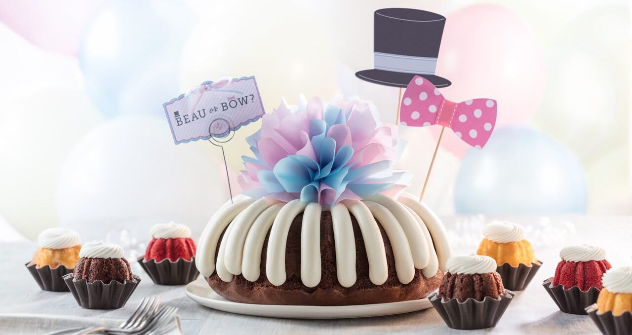 Boy or Girl - Table featuring Bundt Cake with cream cheese frosting, blue and pink bow, paper top hat and pink bow decoration, and beau or bow sign, surrounded by assorted Bundtinis with cream cheese frosting - Shop Now