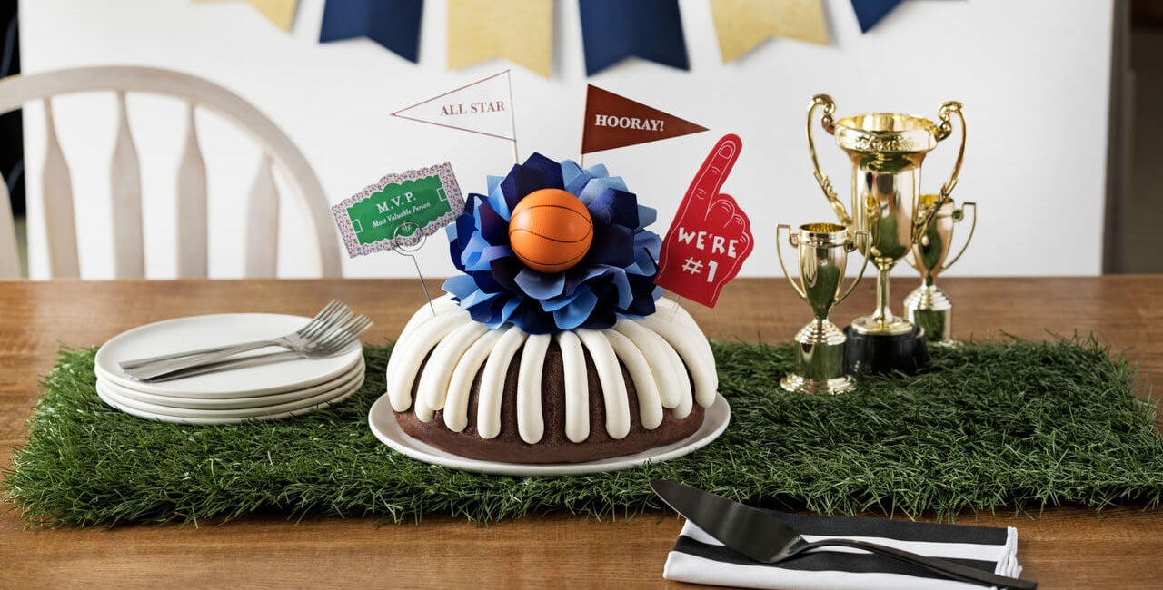 Sports celebration Bundt Cake with cream cheese frosting, blue tissue paper flower, basketball decoration, sports pennants including All Star, Hooray, and We're #1, MVP sign, and gold trophy cups on artificial grass for sports team celebrations and game day parties - Shop Now