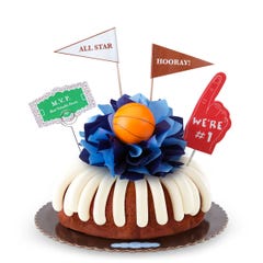 A Carrot bundt cake with frosting, decorated with a mini basketball on top of a bow with different shades of blue in the center of the cake. The cake also contains a We're #1 foam finger, a flag that says All Star and a card that says MVP.