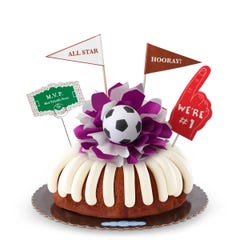 A Carrot bundt cake with frosting, decorated with a mini soccer ball on top of a purple and white bow in the center of the cake. The cake also contains a We're #1 foam finger, a flag that says All Star and a card that says M.V.P.