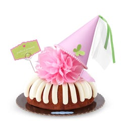 A Carrot bundt cake with frosting, topped with a pink bow and a princess-themed pink party hat with a butterfly on top. A card on the cake reads Prettiest Princess.