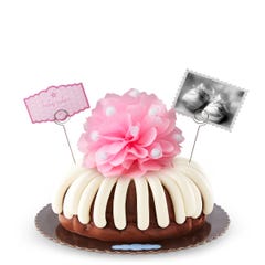 A bundt cake with a pink bow in the center with a small photo card of baby shoes and a pink dotted card that reads baby cakes. Sweet congratulations!