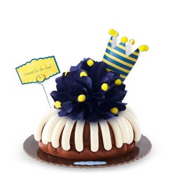 A bundt cake with a navy colored bow with golden pom-poms. A nay and yellow crown tops the cake along with card that says c'round' for the day!.