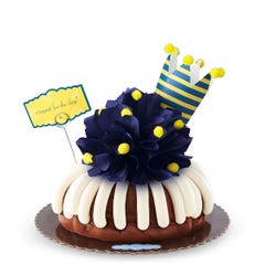 A bundt cake with a navy colored bow with golden pom-poms. A nay and yellow crown tops the cake along with card that says c'round' for the day!.