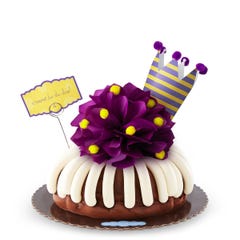 A bundt cake with a plum colored bow with golden pom-poms. A plum and yellow crown tops the cake along with card that says c'round' for the day!.