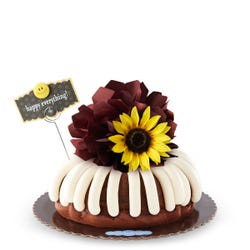 A bundt cake with frosting with a chocolate/brown colored bow and a sunflower topper. There is a decoration card that says happy everything! with a smiley face.