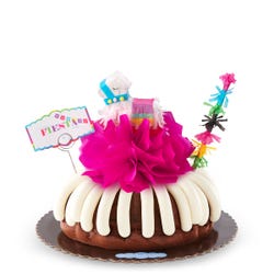 A party-themed bundt cake with a colorful mini piñata at the center and a piñata stick next to it. The cake has a card that reads FIESTA. 