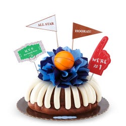 A bundt cake with frosting, decorated with a mini basketball on top of a bow with different shades of blue in the center of the cake. The cake also contains a We're #1 foam finger, a flag that says All Star and a card that says MVP.