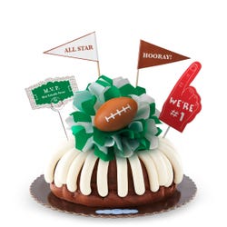 A bundt cake with frosting, decorated with a mini football on top of a green and white bow in the center of the cake. The cake also contains a We're #1 foam finger, a flag that says All Star and a card that says MVP.