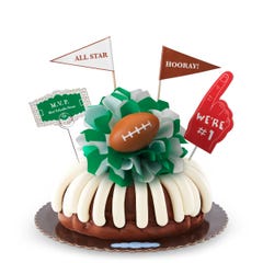A bundt cake with frosting, decorated with a mini football on top of a green and white bow in the center of the cake. The cake also contains a We're #1 foam finger, a flag that says All Star and a card that says MVP.
