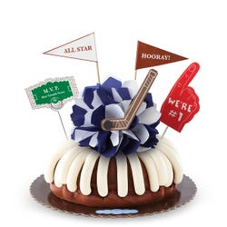 A bundt cake with frosting, decorated with a mini hockey puck on top of a blue and white bow in the center of the cake. The cake also contains a We're #1 foam finger, a flag that says All Star and a card that says M.V.P.