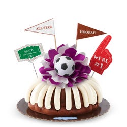 A bundt cake with frosting, decorated with a mini soccer ball on top of a purple and white bow in the center of the cake. The cake also contains a We're #1 foam finger, a flag that says All Star and a card that says M.V.P.