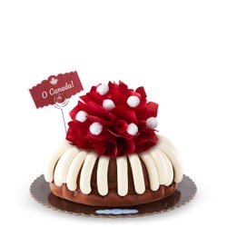 A bundt cake with frosting with the center of the cake topped with a red satin bow with white pom poms. A decorative message card reads O Canada!.