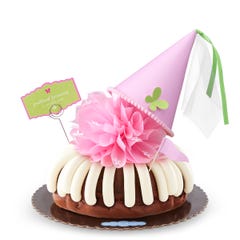 A Chocolate Chocolate Chip bundt cake with frosting, topped with a pink bow and a princess-themed pink party hat with a butterfly on top. A card on the cake reads Prettiest Princess.