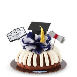 Bundt cake with frosting and topped with a customizable colored bow. It is topped with a diploma rolled up and a graduation cap with a tassel and a card that says you did it!.