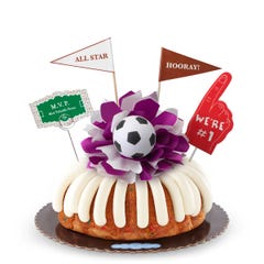 A confetti bundt cake with frosting, decorated with a mini soccer ball on top of a purple and white bow in the center of the cake. The cake also contains a We're #1 foam finger, a flag that says All Star and a card that says M.V.P.