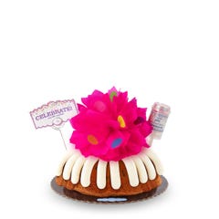 Large decorated bundt cake covered with frosting. Decoration includes a pink bow with confetti sprinkles and a confetti popper with a card that says Celebrate and cue the Gluten Free Chocolate Chip Cookie. 