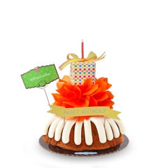 A Gluten Free Chocolate Chip Cookie birthday cake with frosting on top. The cake is decorated with an orange bow and a wrapped gift box with different color dots and topped with a gift bow and a candle. 