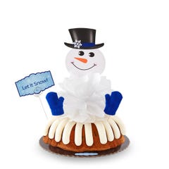 A decorated Gluten Free Chocolate Chip Cookie bundt cake with a snowman with a Gluten Free Chocolate Chip Cookie nose and 2 knit mittens. A card is on the cake that says Let It Snow!.