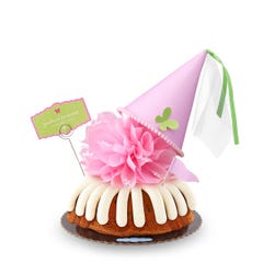 A Gluten-Free Chocolate Chip Cookie bundt cake with frosting, topped with a pink bow and a princess-themed pink party hat with a butterfly on top. A card on the cake reads Prettiest Princess.