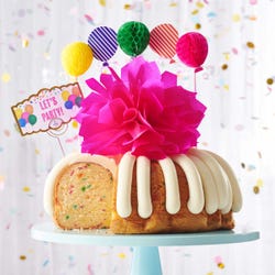 A party-themed bundt cake with colorful 3D balloons. The cake has a card that reads LET'S PARTY. 