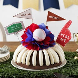 A bundt cake with frosting, decorated with a baseball on top of a purple and white bow in the center of the cake. The cake also contains a We're #1 foam finger, a flag that says All Star and a card that says M.V.P.