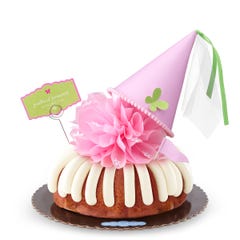 A Strawberries & Cream bundt cake with frosting, topped with a pink bow and a princess-themed pink party hat with a butterfly on top. A card on the cake reads Prettiest Princess.