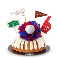 A Vanilla bundt cake with frosting, decorated with a baseball on top of a purple and white bow in the center of the cake. The cake also contains a We're #1 foam finger, a flag that says All Star and a card that says M.V.P.