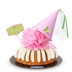A Vanilla bundt cake with frosting, topped with a pink bow and a princess-themed pink party hat with a butterfly on top. A card on the cake reads Prettiest Princess.