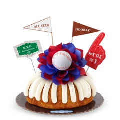 A White Chocolate Raspberry bundt cake with frosting, decorated with a baseball on top of a purple and white bow in the center of the cake. The cake also contains a We're #1 foam finger, a flag that says All Star and a card that says M.V.P.